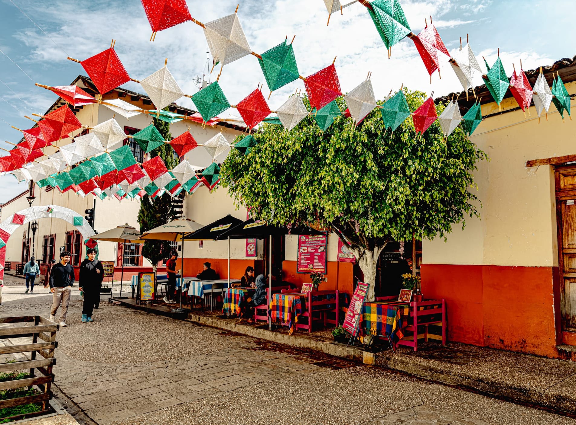 Digital nomad life in San Cristóbal de las Casas Mexico, outdoor café and restaurant scene on walkable street, colorful papel picado decorations overhead, everyday street life in the Chiapas Highlands, cafés for digital nomads and remote workers, relaxed dining and working atmosphere in southern Mexico