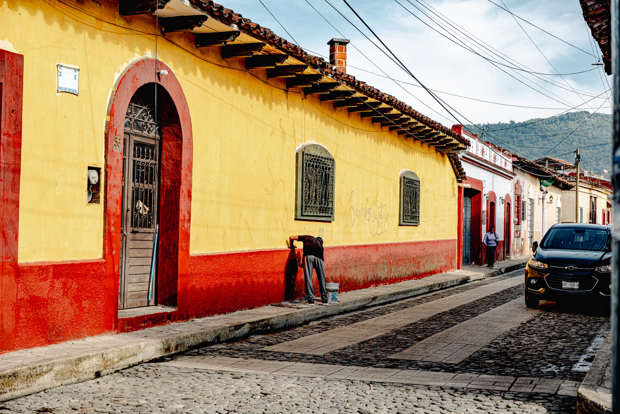 Digital nomad life in San Cristóbal de las Casas Mexico, colorful colonial street with yellow and red walls, person painting building facade on walkable cobblestone street, everyday maintenance and care in historic neighborhood, lived in city life in the Chiapas Highlands, daily rhythms experienced by digital nomads and remote workers in southern Mexico