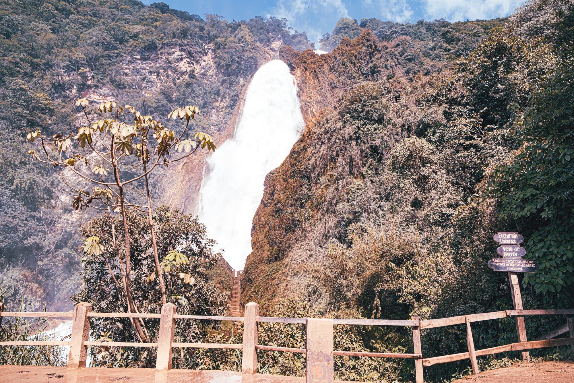 Velo de Novia waterfall at El Chiflón near San Cristóbal de Las Casas, Chiapas, Mexico, viewed from the lower lookout with wooden railings and trail signage, showing the main waterfall cascading through dense jungle at the El Chiflón waterfalls park, a popular day trip often combined with Montebello Lakes
