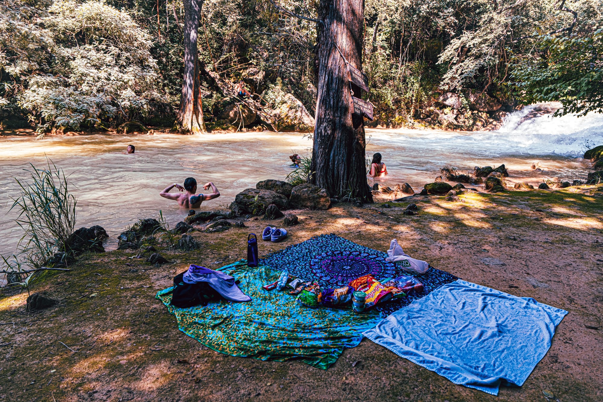 People swimming and relaxing along the river at El Chiflón waterfalls near San Cristóbal de Las Casas, Chiapas, Mexico, with picnic blankets and personal items set up beside the water at a designated swimming area within the El Chiflón waterfall park, a popular day trip often combined with Montebello Lakes