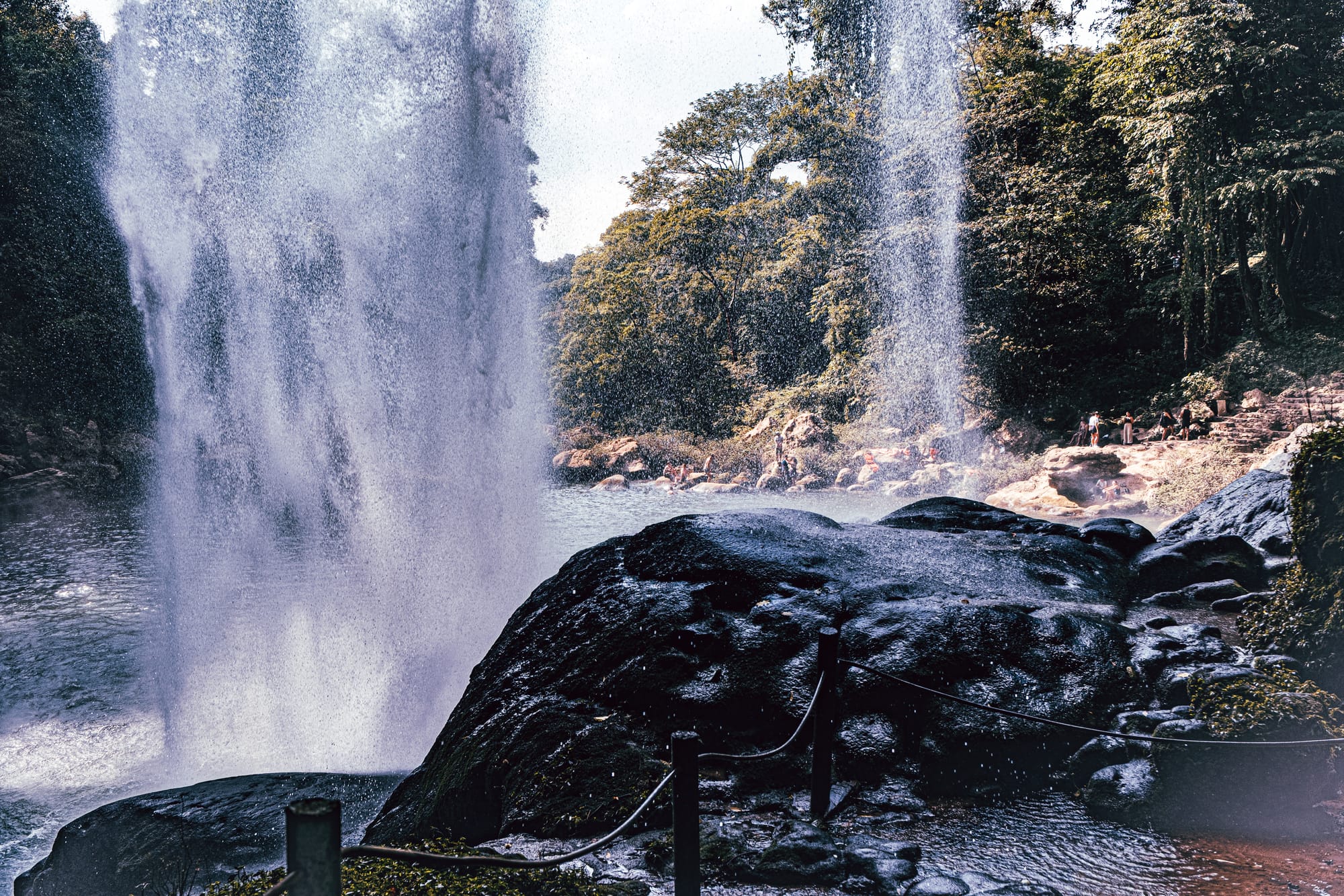 View from behind Misol-Há waterfall near Palenque, Chiapas, looking out toward the natural pool and surrounding jungle, with water cascading in the foreground and visitors gathered along the rocky edges, capturing the perspective from inside the waterfall cave area at one of the most visited waterfalls near Palenque in southern Mexico