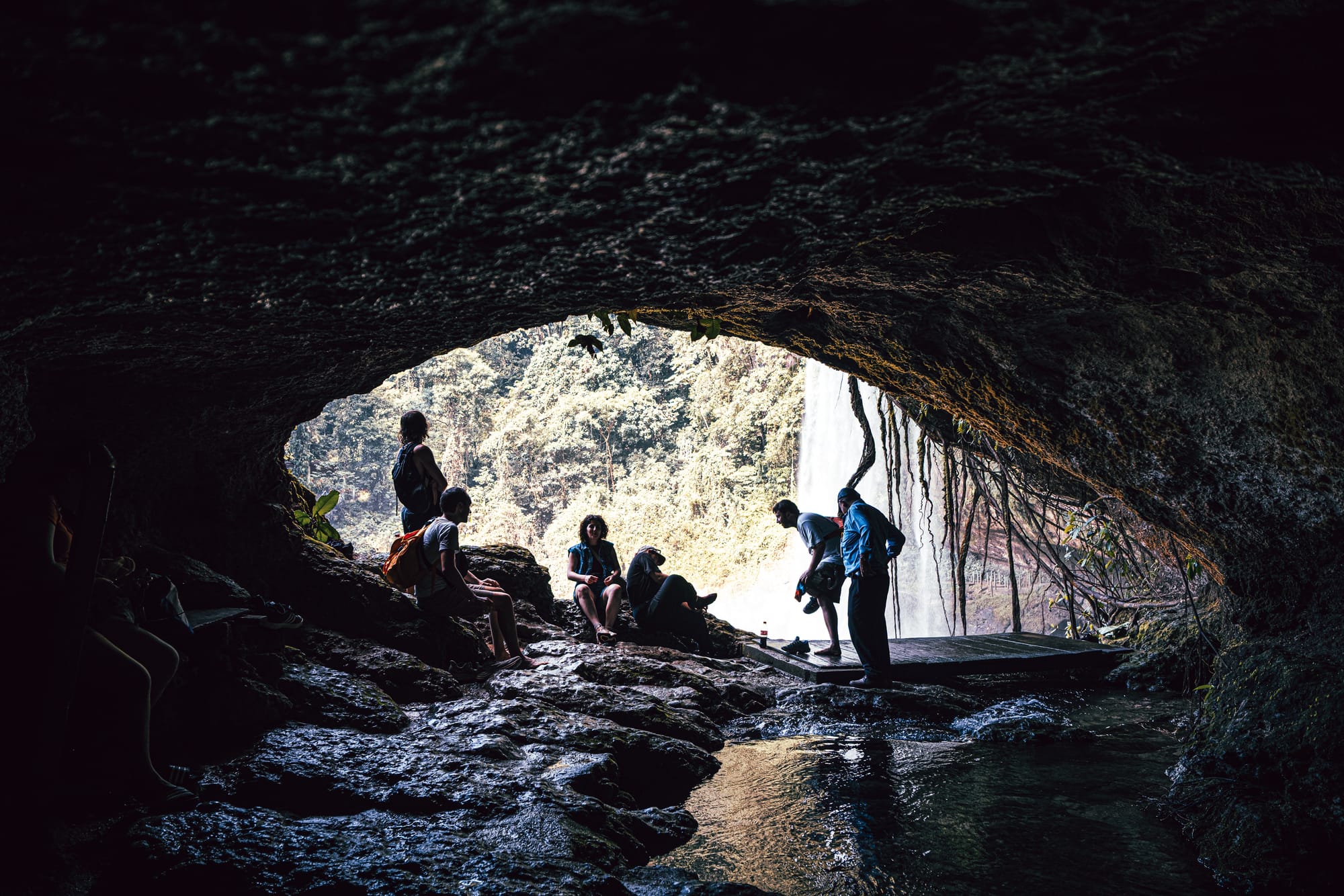View from inside the cave behind Misol-Há waterfall near Palenque, Chiapas, showing visitors resting and standing beneath a rocky overhang with shallow water flowing through the cave, framed by hanging roots and the waterfall visible at the cave entrance, highlighting the walk-behind waterfall experience at one of the most popular waterfalls near Palenque in southern Mexico