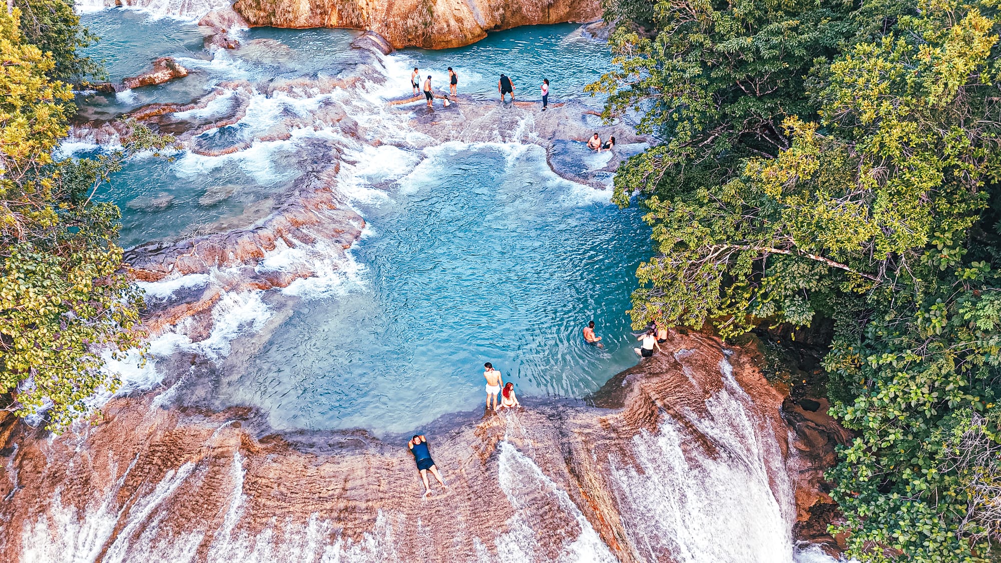 Roberto Barrios waterfalls near Palenque, Chiapas, viewed from above showing natural turquoise pools formed by cascading water over smooth limestone terraces, with visitors swimming, relaxing, and standing along the rock edges, illustrating the expansive, multi-tiered waterfall system that makes Roberto Barrios one of the most impressive waterfalls near Palenque in southern Mexico
