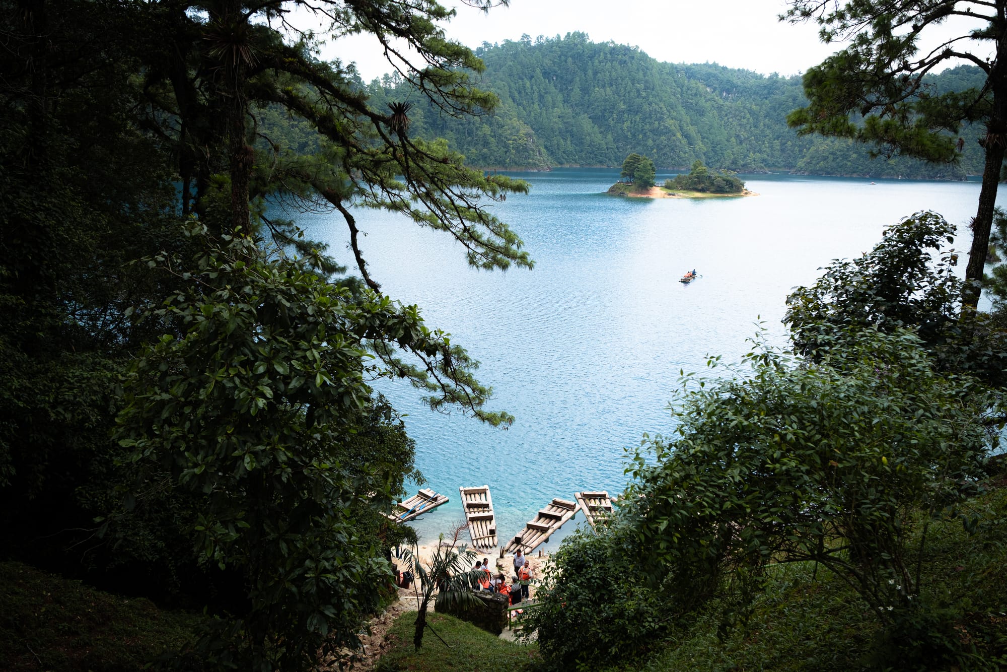 View of Montebello Lakes in Chiapas from a forested hillside, showing wooden rafts on the shore, turquoise water, and a small island beneath the surrounding pine-covered hills