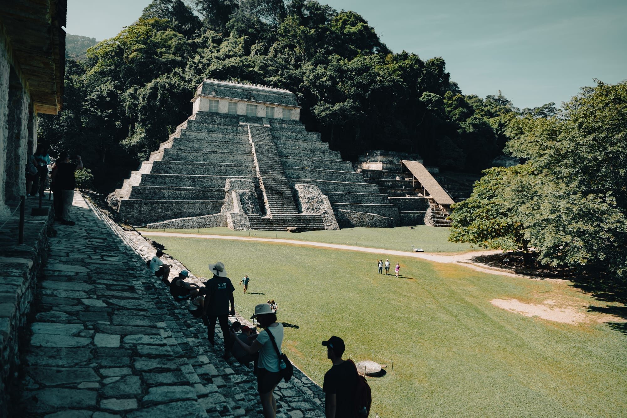 Palenque Mayan ruins Temple of the Inscriptions viewed from the plaza, stepped pyramid rising above grassy courtyard in Chiapas Mexico, visitors walking across Palenque archaeological site, iconic Maya temple near San Cristóbal de las Casas surrounded by jungle
