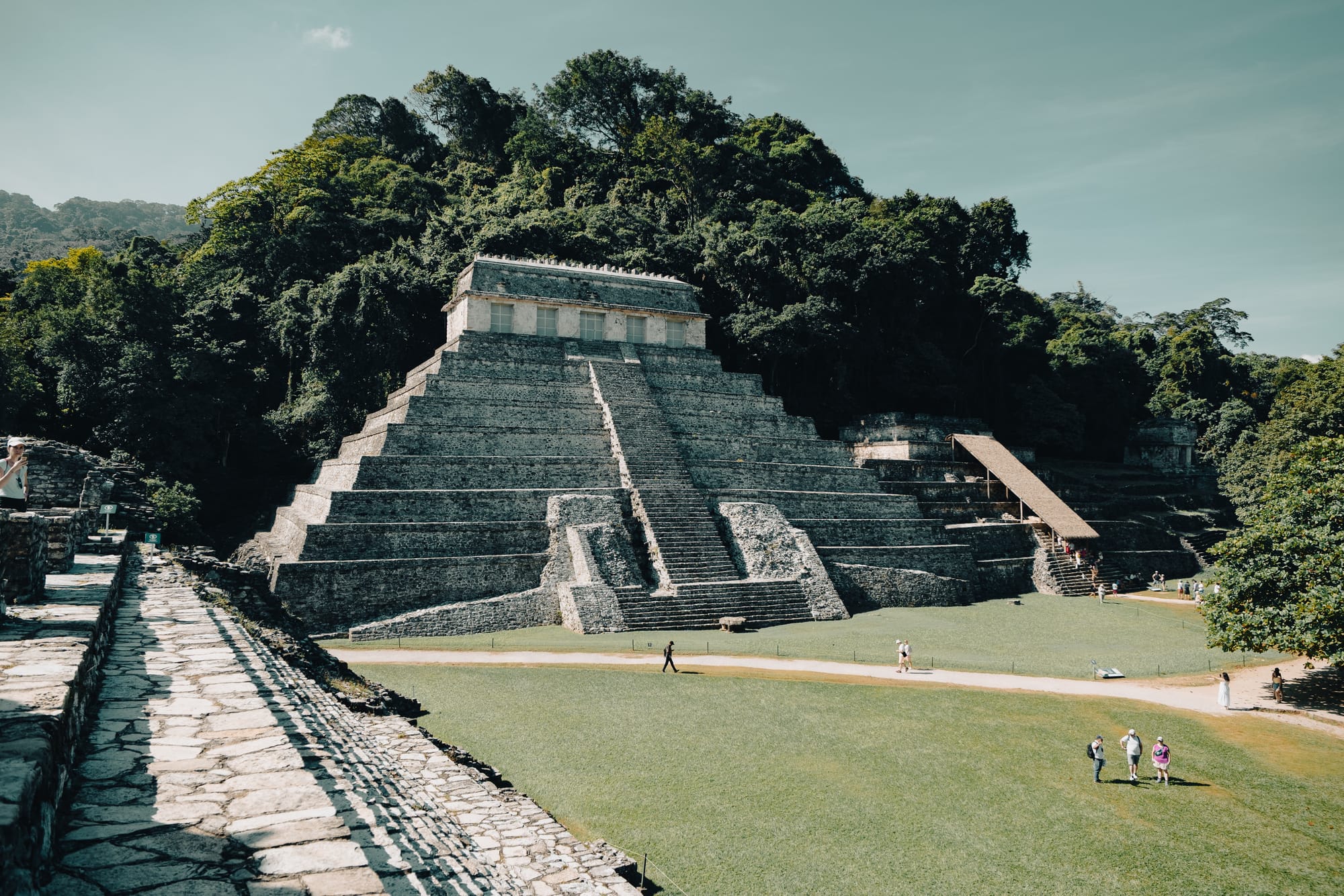 Temple of the Inscriptions at Palenque Mayan ruins viewed from elevated stone walkway, large stepped pyramid dominating central plaza at Palenque archaeological site in Chiapas Mexico, iconic Maya temple surrounded by jungle near San Cristóbal de las Casas, UNESCO World Heritage Mayan ruins with visitors walking across grassy courtyard