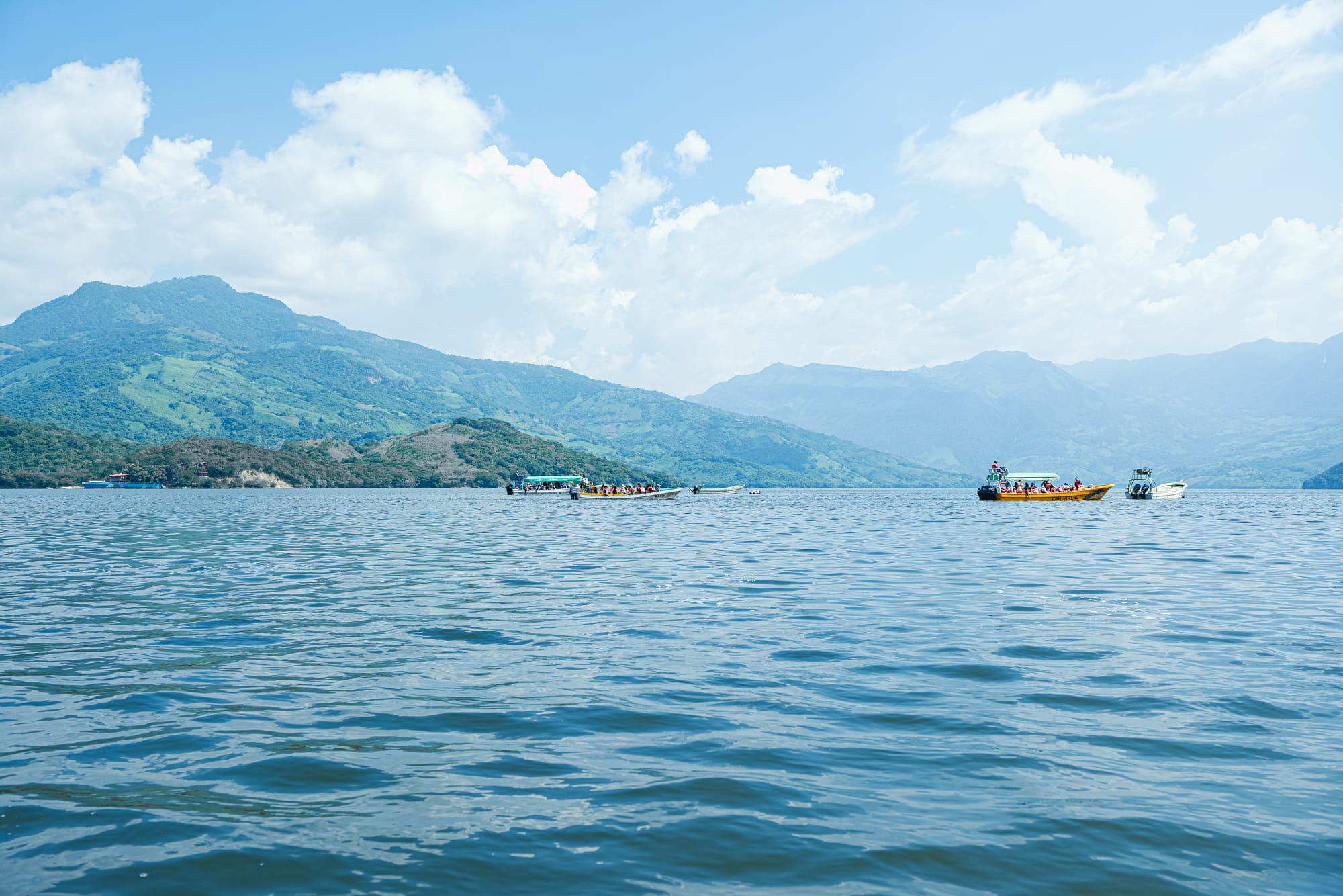 Small tour boats on the Grijalva River near the entrance to Sumidero Canyon, with rolling green hills and mountains in the background on a clear day