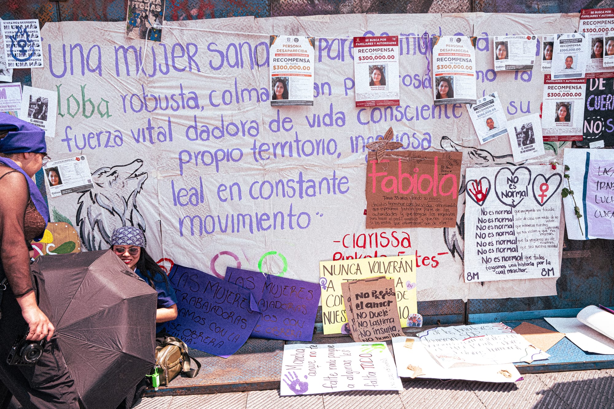 Women sitting beside a wall of feminist posters and missing-person flyers during the 8M march in Mexico City calling for justice for victims of femicide